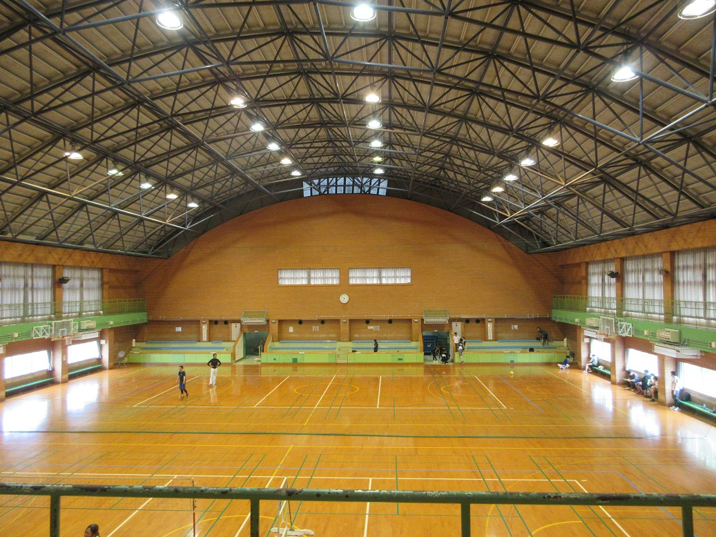むき出しの鉄骨トラス構造の天井に多数の照明が灯り、オレンジ色の壁と木製のフローリングが広がる体育館の内部で、数人の利用者がコート上で活動したり壁際で休憩したりしている様子を俯瞰で捉えた写真