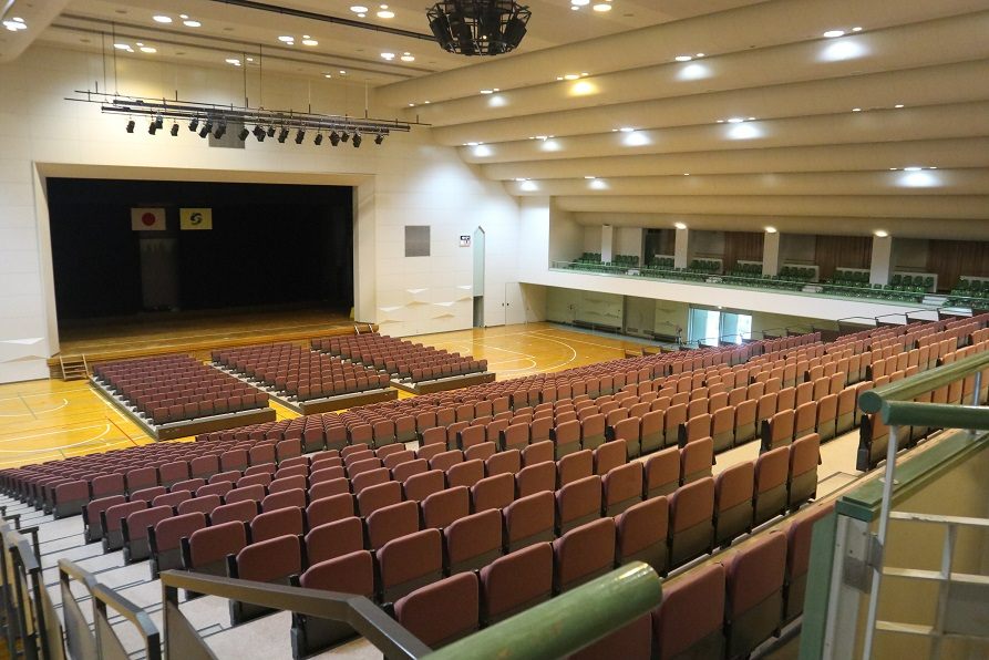 広々とした多目的ホール内を後方の観客席から見下ろした全景で、手前には茶色の座席が並ぶ巨大な固定式スタンドがあり、その先には板張りのフロアとステージ、そして天井に設置された多数の照明設備が映し出されている移動席の写真