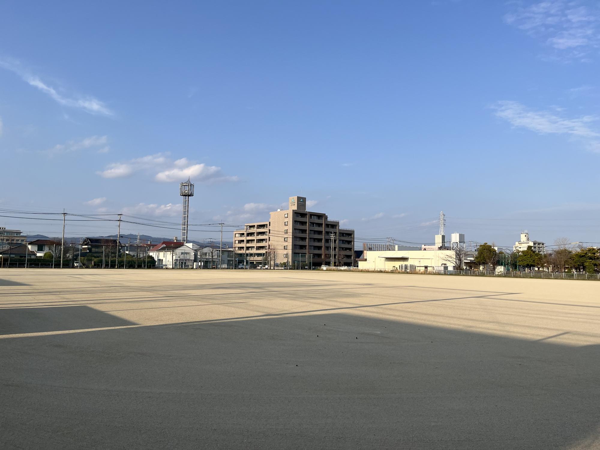 青空の下に広がる広大な砂地のグラウンドの先に、茶色のマンションや住宅街、電波塔、電柱などが立ち並び、手前には建物による大きな影が落ちている、西神野運動広場の写真