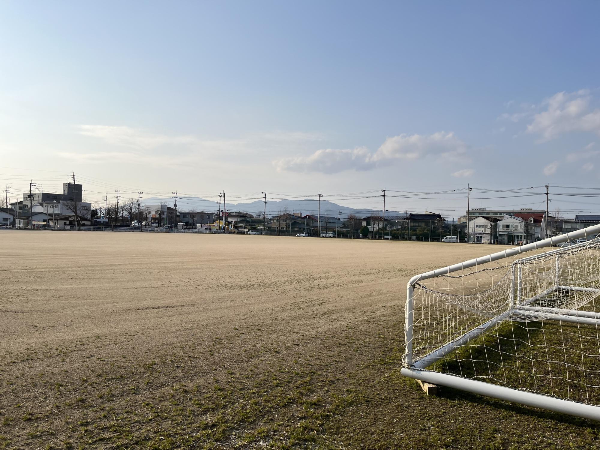 晴れ渡る空の下に広がる土のグラウンドの右端に白いサッカーゴールの一部が映り込み、遠景には並んだ住宅街や電柱の列、そして微かに霞む山並みが地平線に沿って続く、西神野運動広場の写真