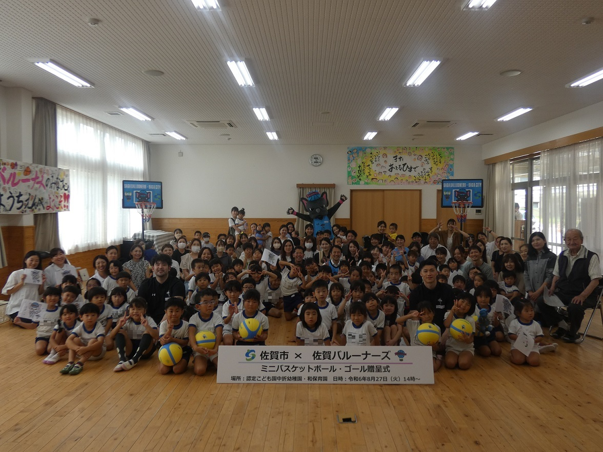 中折幼稚園での贈呈式での子供たちと大人、バルーナーズの選手が集まって座り、その後ろにミニバスケットゴールつと両手を広げたバルたんが立っている集合写真
