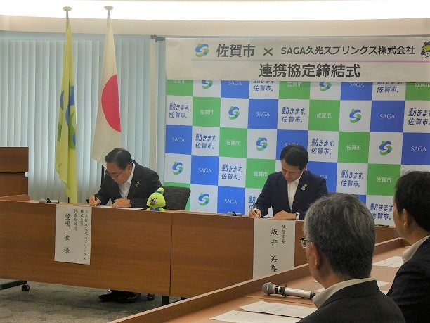 佐賀市と企業の連携協定締結式で関係者が国旗と社旗の前で書類に署名する様子を捉えた写真