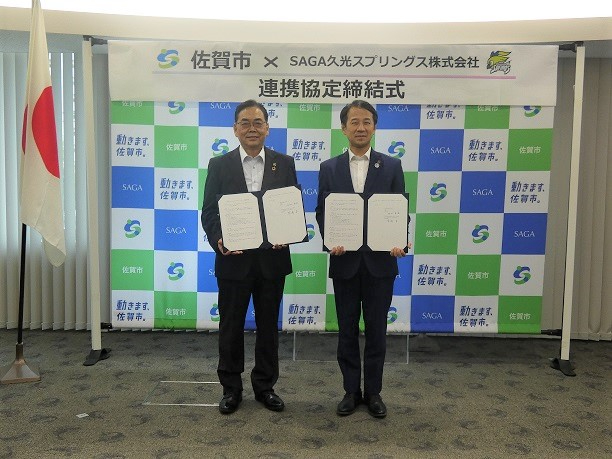 佐賀市と企業の連携協定締結式で二人の関係者が並んで協定書を掲げ背景のロゴ壁面前に立つ写真