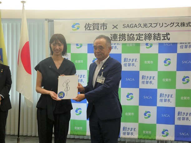 佐賀市と企業の連携協定締結式で二人が石井優希選手のサイン色紙を共に持ち背景のロゴ壁面前に立つ写真