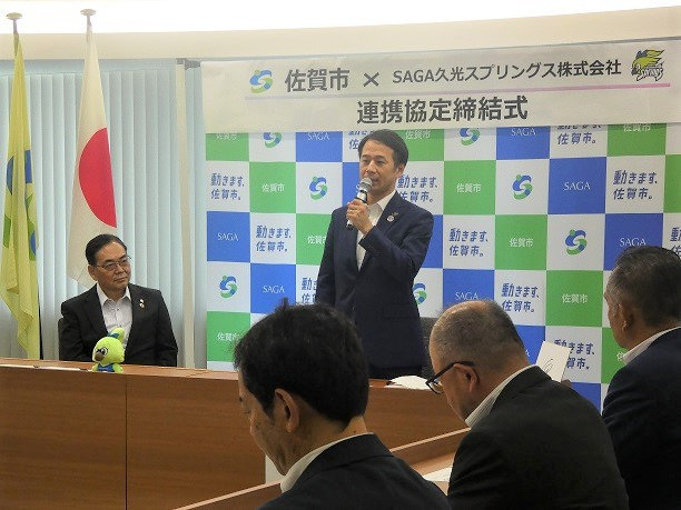 佐賀市と企業の連携協定締結式で坂井英隆市長が国旗を背にマイクで挨拶し出席者が注目する様子を捉えた写真