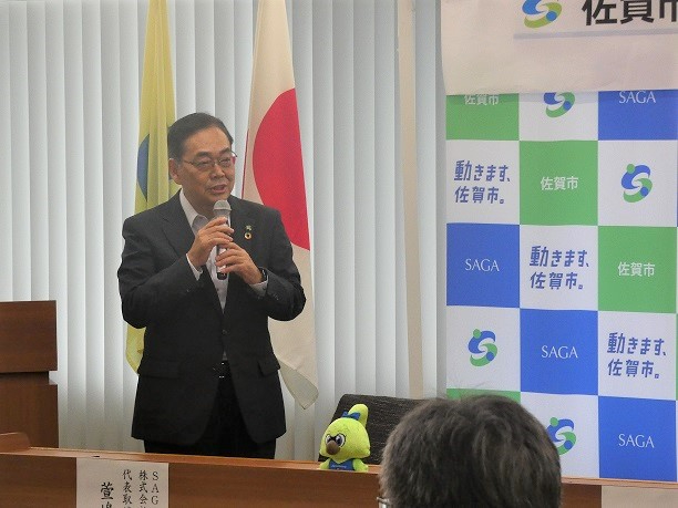 日本旗と市旗を背にスーツ姿の萱嶋章代表取締役が壇上でマイクを手に挨拶する様子を捉えた写真挨拶