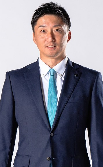 濃紺のスーツに白いシャツと明るい青のネクタイを身につけた宮永雄太ヘッドコーチ兼ゼネラルマネージャーがシンプルな背景の前で直立する姿を写した写真