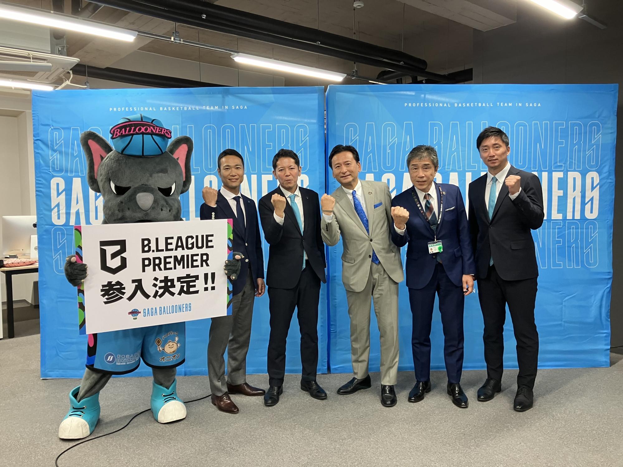 佐賀バルーナーズのマスコットキャラクターのバルたんが「B.LEAGUE PREMIER参入決定！！」と書かれたボードを掲げ、スーツ姿の男性5名と共に青い背景の前でガッツポーズをして参入を祝っている、発表会の様子の写真