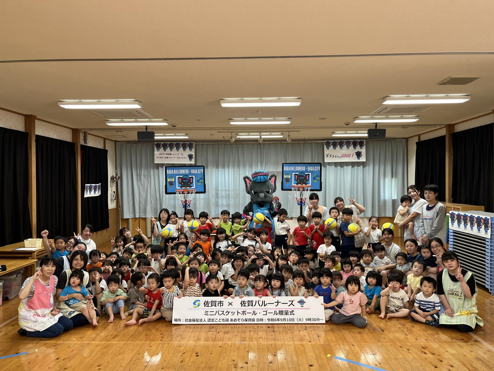 あおぞら保育園での贈呈式で、子供たちや園の先生が集まって座っている後ろにミニバスケットゴール2つとバルたんが立っている集合写真