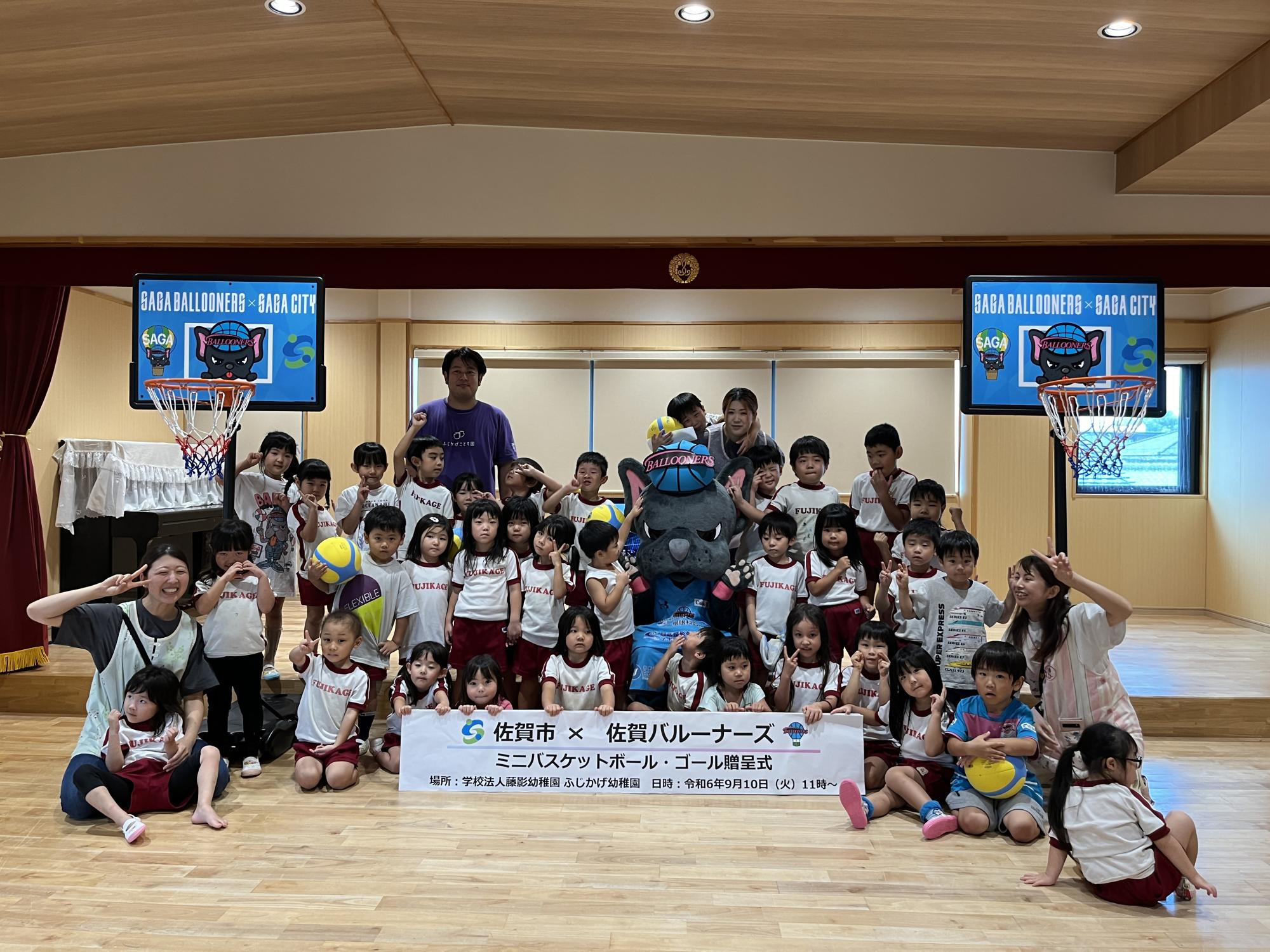 ふじかげこども園での贈呈式で、子供たちや先生が集まっている中央にバルたんが座り、両脇にはミニバスケットゴール2つが置かれている集合写真