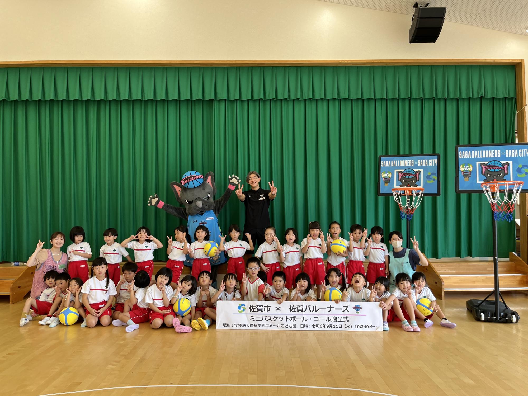 エミールこども園での贈呈式で、子供たちが2列に並んだ後ろにピースをする佐賀バルーナーズの選手と両手を広げたバルたんが立ち、右に2つのミニバスケットゴールが置かれている集合写真
