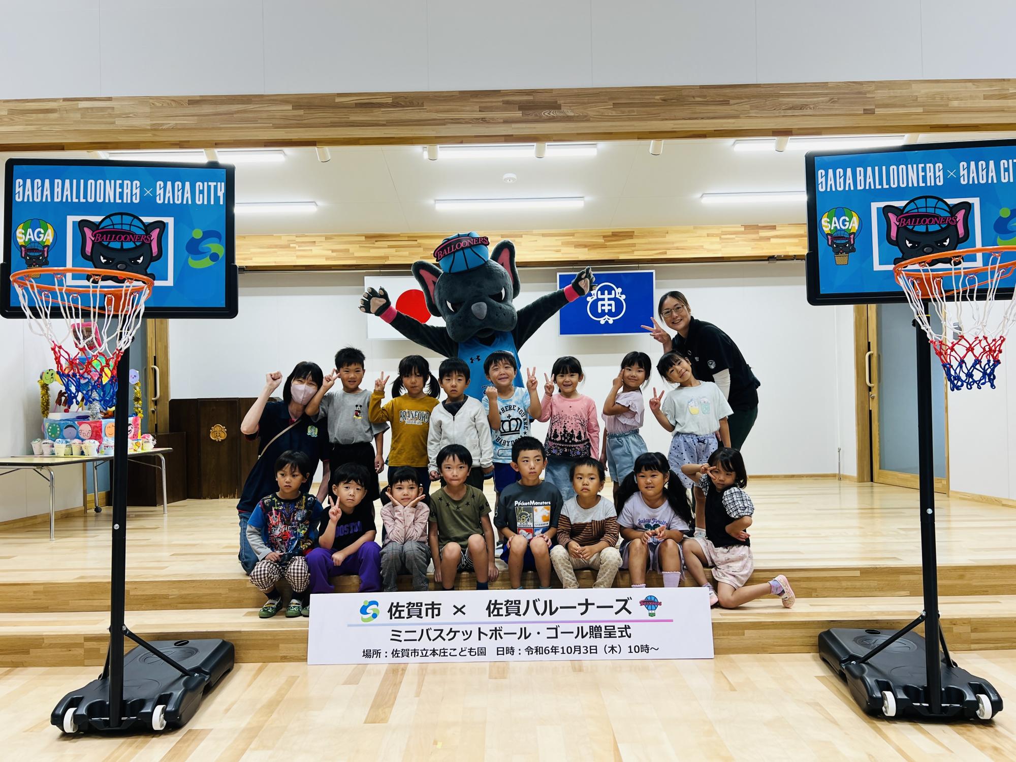本庄こども園での贈呈式で、2列に並んだ子供たちの後ろに両手を広げたバルたんが立ち、その前方両サイドにミニバスケットゴールが置かれている集合写真