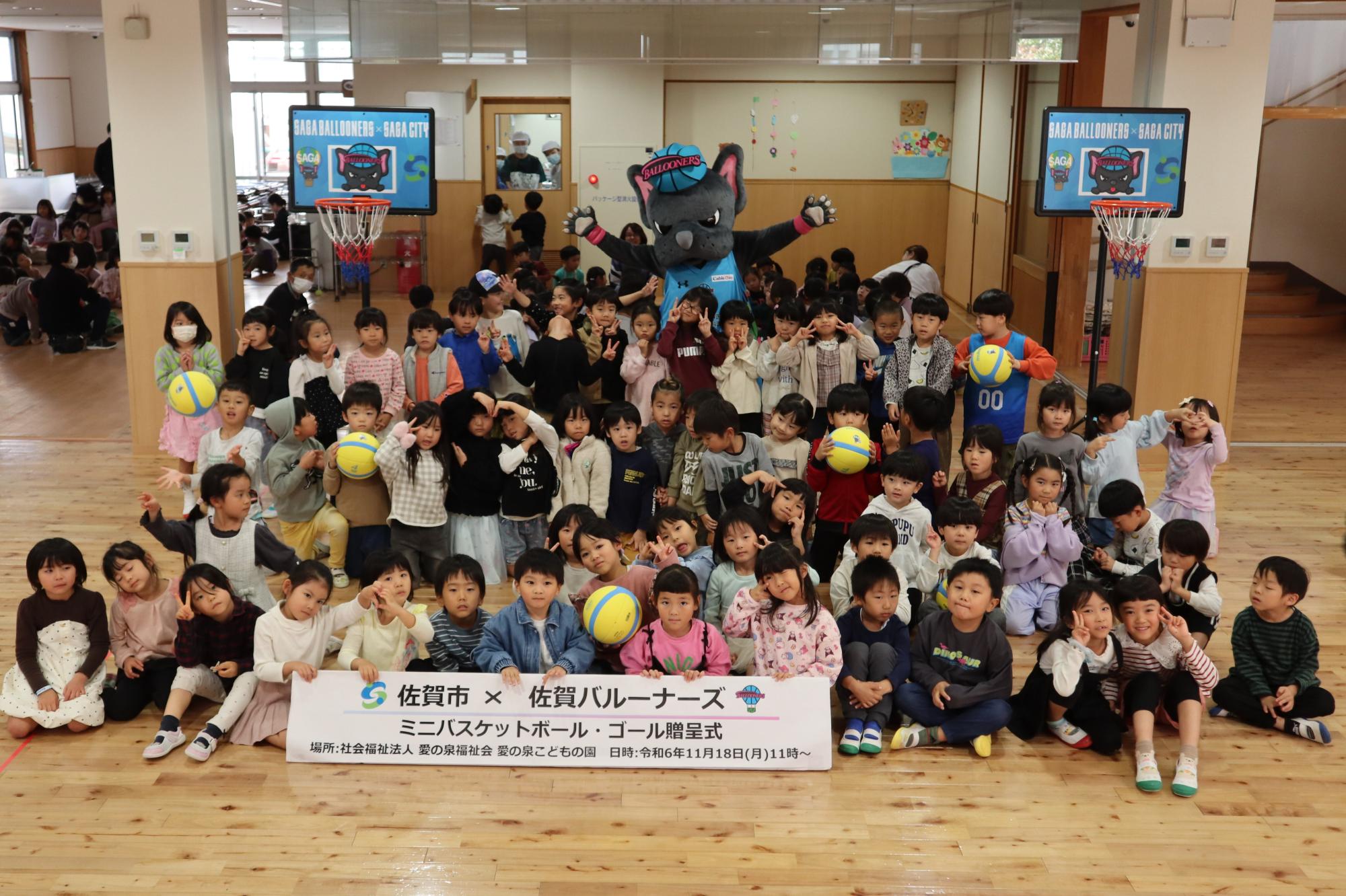 愛の泉こどもの園での贈呈式で、たくさんの子供たちが並んだ後ろで両手を広げたバルたんが立ち、その両サイドにミニバスケットゴールが置かれている集合写真