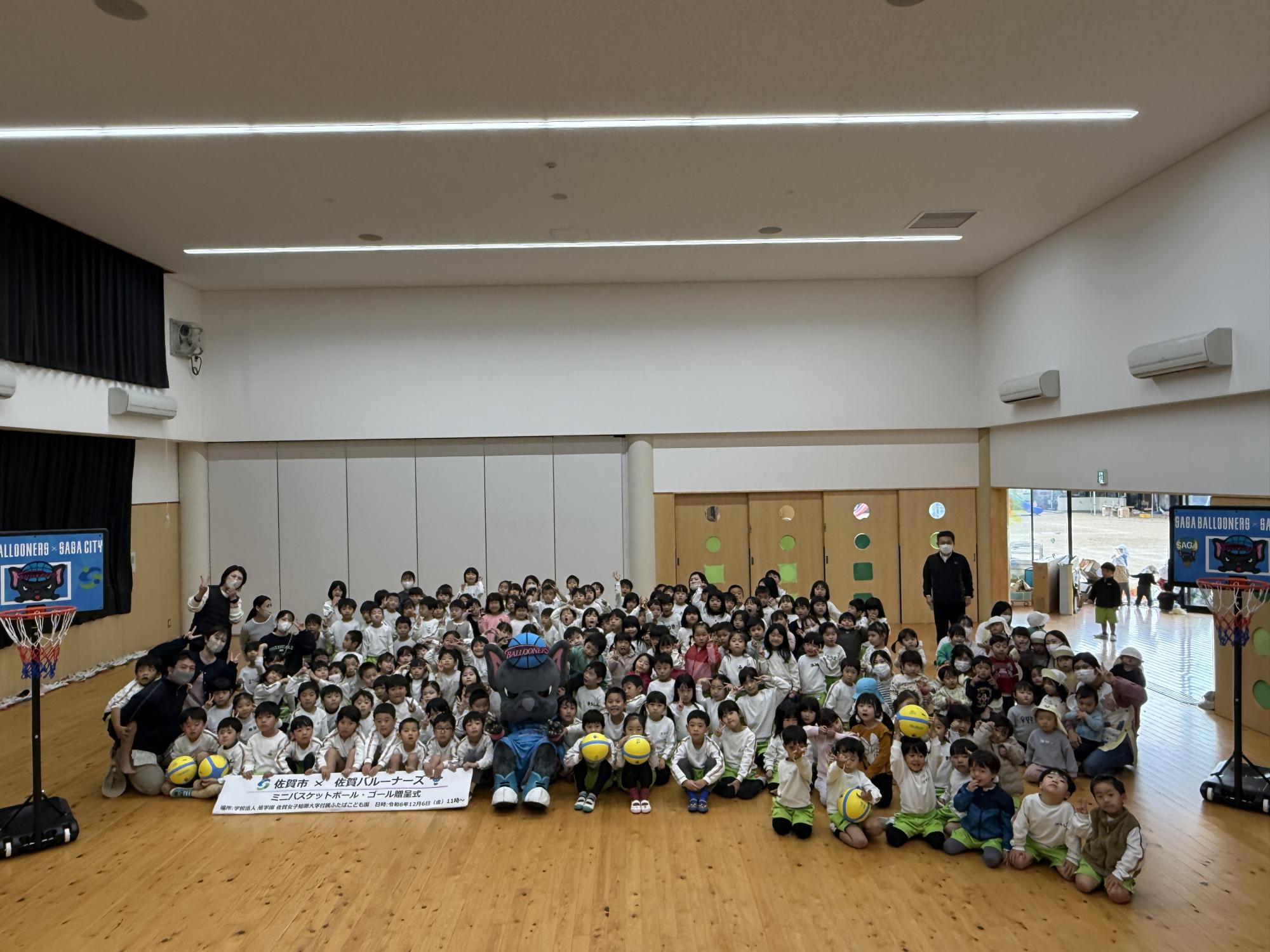 佐女短附属ふたばこども園での贈呈式で、たくさんの園児がならんだ中央にバルたんが座り、両サイドにミニバスケットゴールが置かれている集合写真