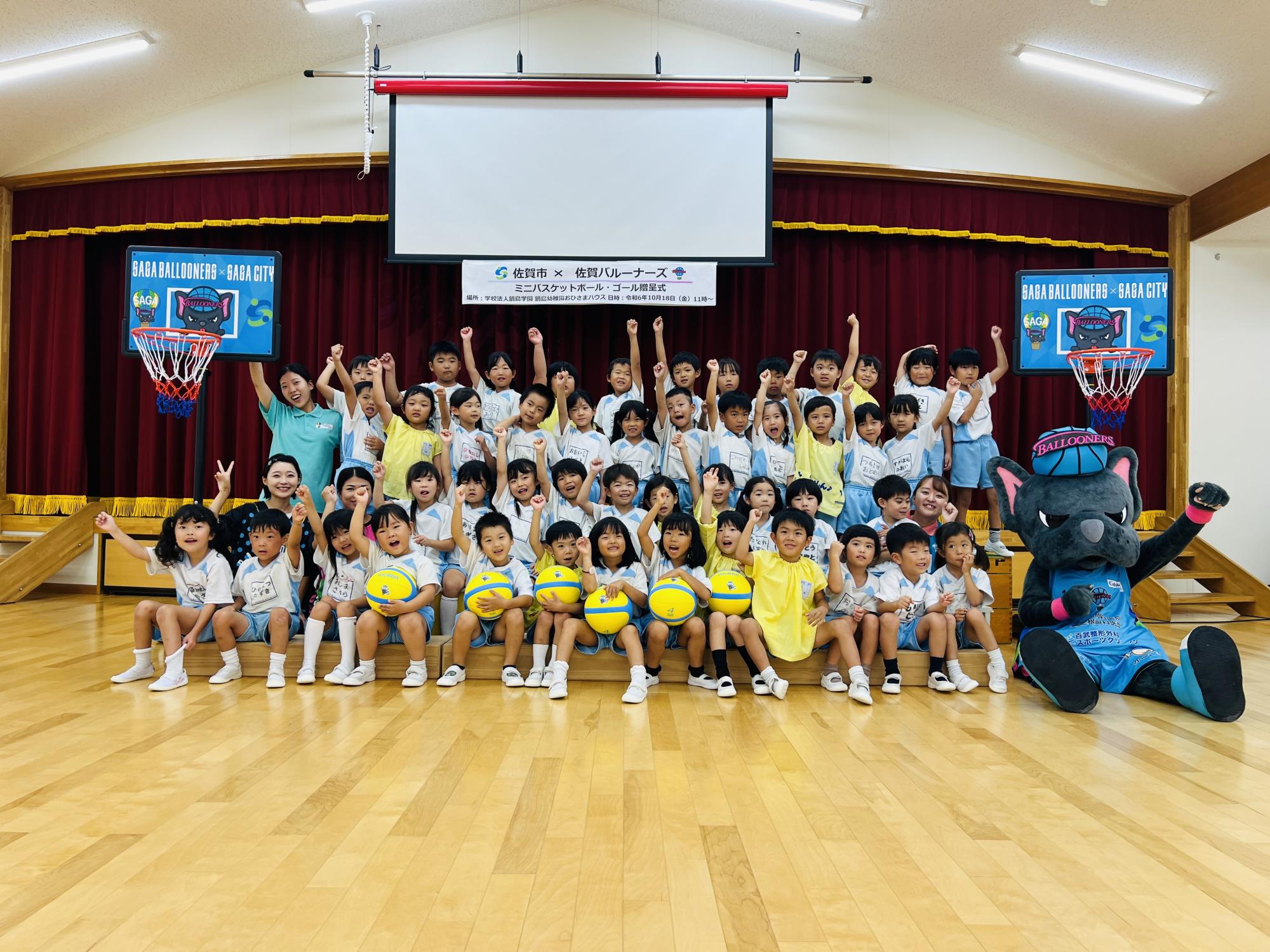 鍋島幼稚園おひさまハウスでの贈呈式で、並んで手を上に挙げるポーズをしている子供たちと、前列右側に座っているバルたんの集合写真