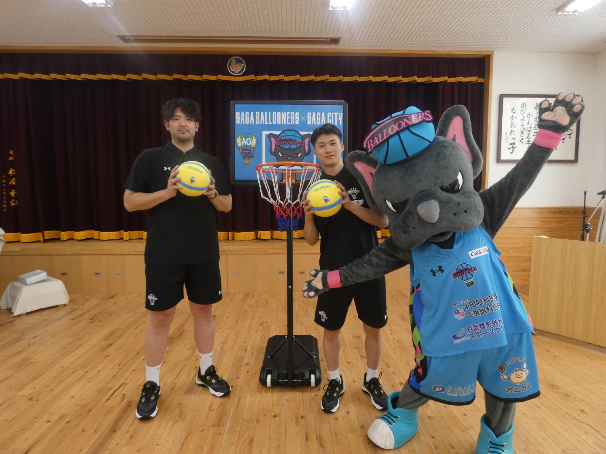 佐賀バルーナーズの選手2人がバスケットボールを持ち、マスコットキャラクターのバルたんが両手を広げ、ミニバスケットゴールの前に並んで立っている写真