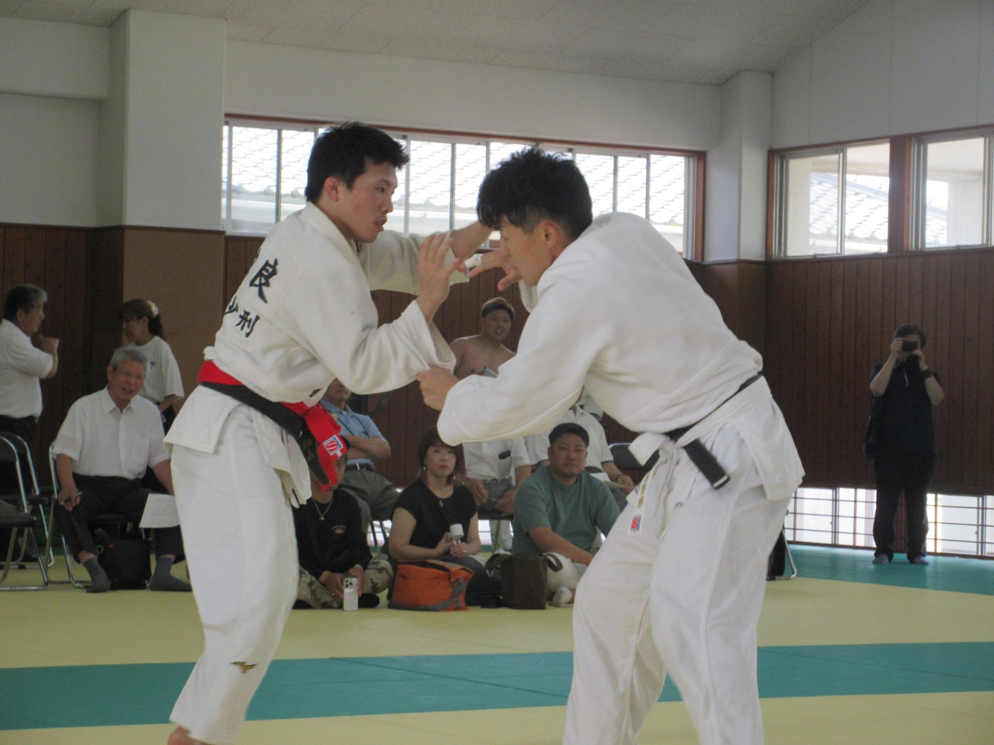 明るい道場で、白の柔道着を纏った二人の選手が互いに襟や袖を掴み合い、真剣な表情で技を掛けようと激しく組み合う様子を、背後の席で静かに見守る観客たちと共に捉えた、緊張感漂う柔道の試合風景を写した写真