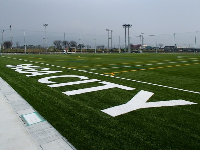 曇り空の下に広がる鮮やかな緑の人工芝サッカーコートのサイドライン際に、白く大きな文字で「SAGA CITY」と印字された様子を近距離から捉えた、周囲に照明塔が立ち並ぶスポーツ施設の屋外写真