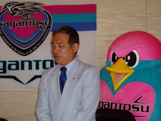 サガン鳥栖の大きなロゴ旗とマスコットキャラクターのウィントスを背景に、水色のスーツに青いネクタイを締めた松本監督が、真剣な面持ちで斜め下を見つめながら記者会見で発言している様子を横から捉えた写真