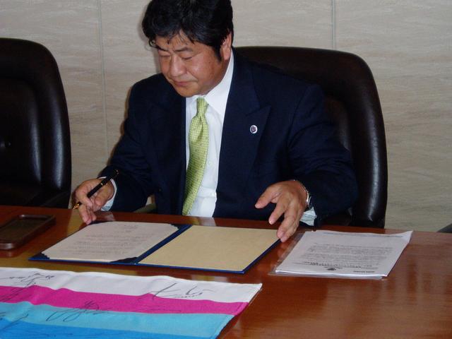 紺色のスーツに黄緑色のネクタイを締めた井川社長が、木製のテーブルで青いバインダーに挟まれた書類を前にペンを握り、真剣な表情で内容を確認しながら署名を行おうとしている場面を正面から捉えた写真