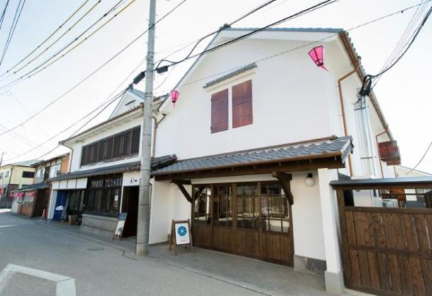 白壁が美しく修復された伝統的な木造建築の街並みにおいて、手前の建物には木製の格子戸や看板が設置され、軒下にはピンク色の提灯が吊るされた情緒ある昼間の風景を、道路沿いから見上げるような角度で捉えた、旧久富家（整備後）の写真