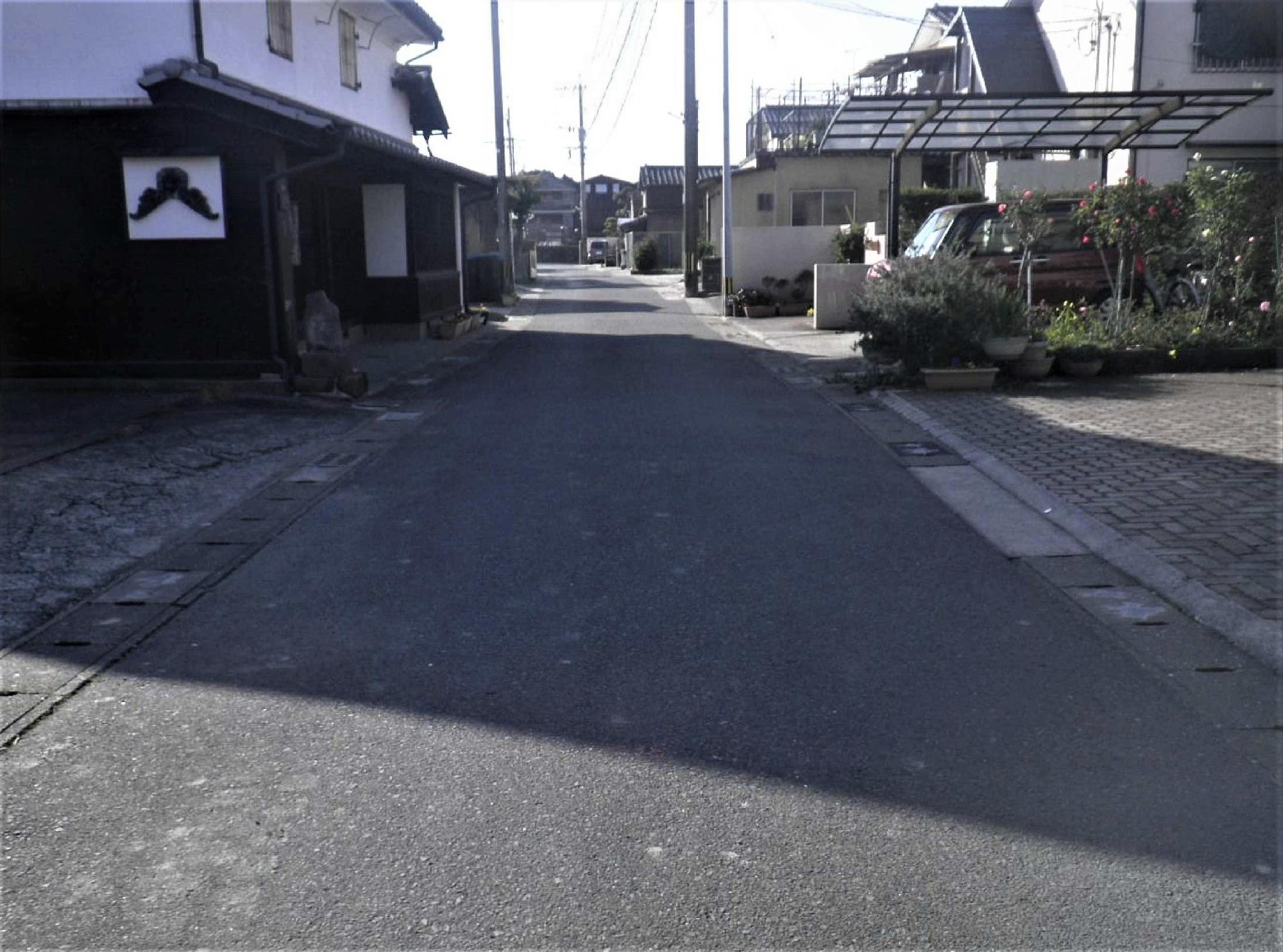 左側に白壁の伝統的な蔵のような建物が並び、右側には現代的な住宅やカーポート、植木鉢が置かれた生活感のある風景が広がる、奥へとまっすぐ続く舗装された静かな路地を逆光気味の落ち着いたトーンで捉えた、長崎街道再整備（整備前平成30年）の写真