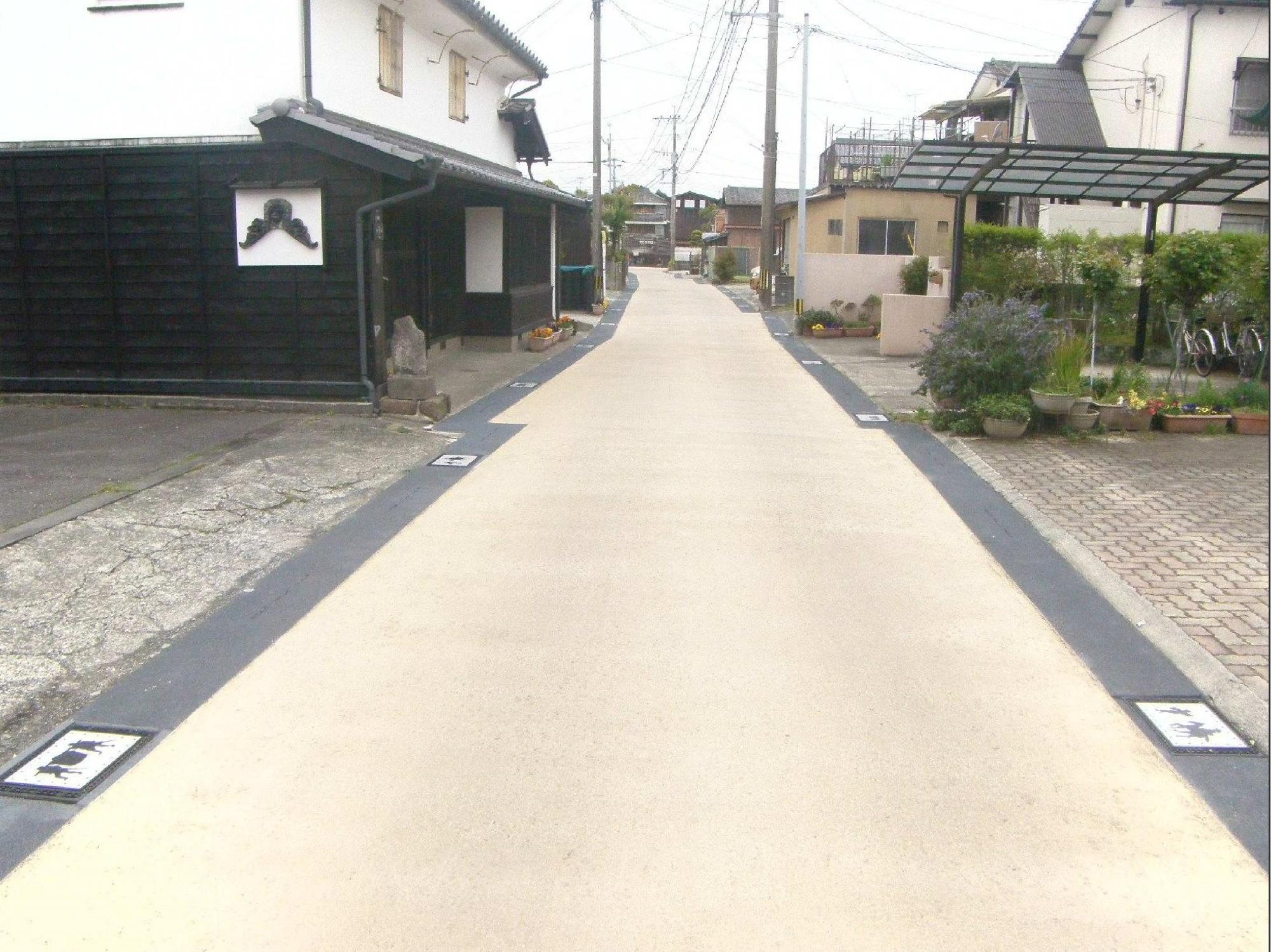 左側に白壁の蔵造りの建物、右側に現代的な住宅やカーポートが並ぶ路地において、路面が明るいベージュ色に舗装され、両端の黒い境界部分には等間隔で意匠を凝らしたタイルが埋め込まれた、歴史と現代が共存する長崎街道再整備（整備前平成31年）の写真