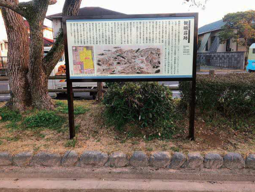 左側に太い幹の古木が立ち、その傍らに「観頤荘跡」と記された歴史解説板が設置されており、解説板には当時の様子を描いた古地図や詳細な説明文が掲載され、背後の住宅街の風景と共に史跡の場所を静かに示す観頤荘跡（整備後）の写真