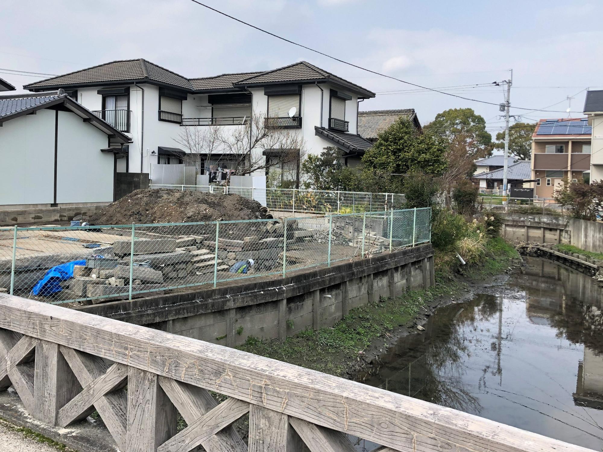 手前の木目調の欄干がある橋の向こう側に、コンクリートの護岸で整備された川が流れ、対岸のフェンスで囲われた空き地には土砂や瓦礫が積まれており、その背景に現代的な白い二階建ての住宅が立ち並ぶ、住宅街の工事現場と水辺の風景を捉えた、思案橋（整備前）の写真