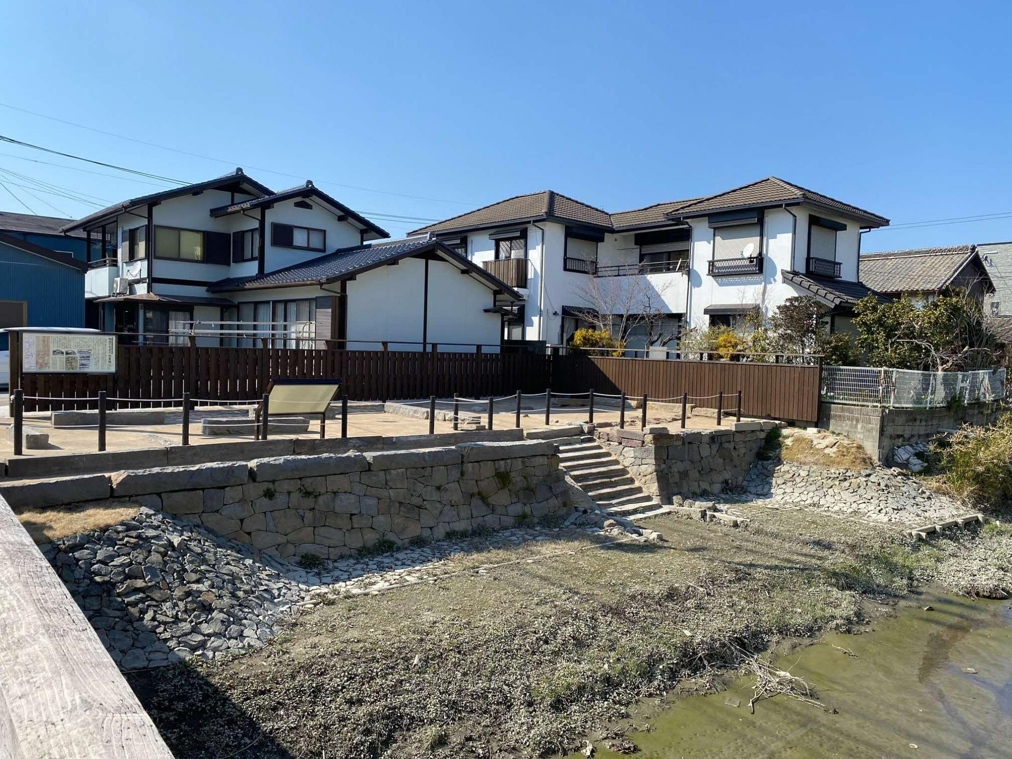 手前の橋から見て川の対岸に、美しく積み直された石垣と川面へ降りる石階段が整備され、その上の敷地には歴史解説板や木製の柵、ベンチが配置されて史跡公園のように整えられた、背景の住宅街と共に歴史を伝える水辺の風景を捉えた、思案橋（整備後）の写真