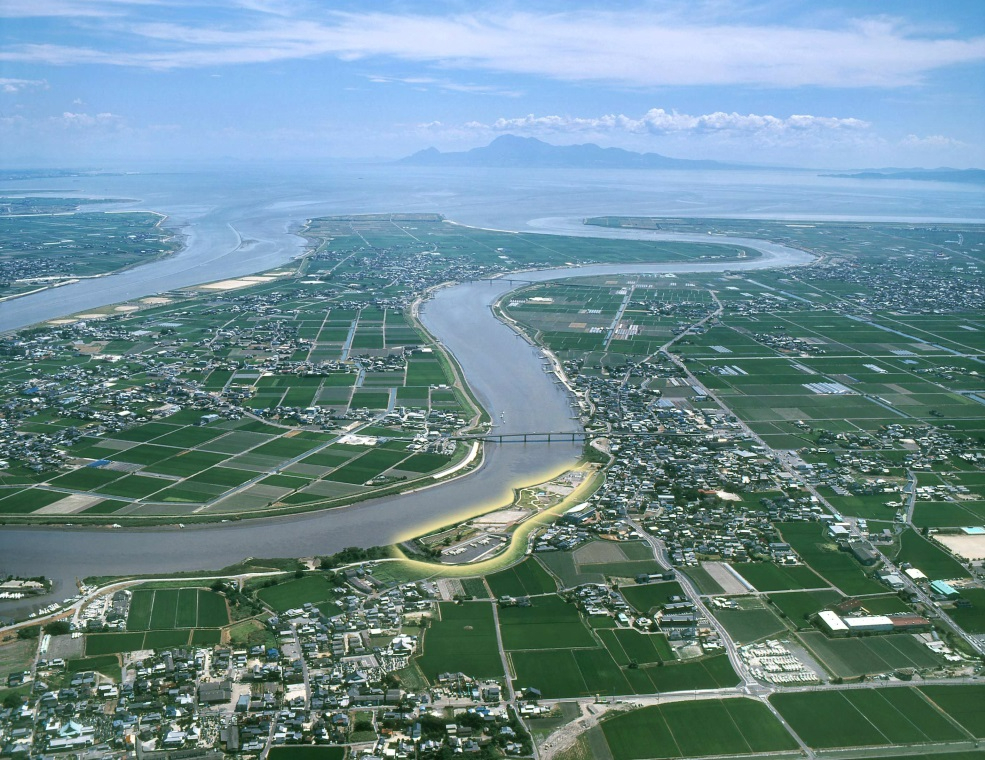 広大な田園地帯の中をゆったりと流れる大きな川の湾曲部に沿って、三重津海軍所跡を含むエリアが光る線で囲まれて強調されており、遠くには海と山影が広がる、歴史的遺構と周辺環境の位置関係を空から俯瞰で捉えた写真