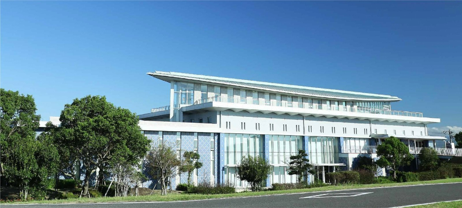 青空を背景に、緩やかな曲線を描く大きな屋根が特徴的な、ガラス張りの開放的な外観を持つ「佐野常民と三重津海軍所跡の歴史館」の全景を、手前の緑豊かな木々や道路と共に捉えた、現代的で清々しい建築デザインが際立つ写真