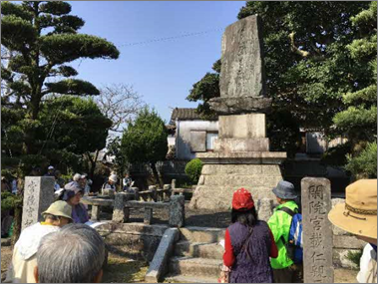 松林や生垣に囲まれた屋外の史跡において、中央に立つ大きな石碑を囲むように、帽子を被った複数の見学者が熱心に碑文を読んだりメモを取ったりしている、佐野常民生誕記念碑の写真