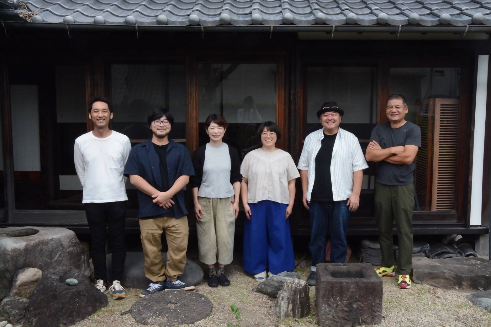趣のある日本家屋の瓦屋根と縁側を背景に、メンバー男女6人が和やかな表情で横一列に並んだ集合写真