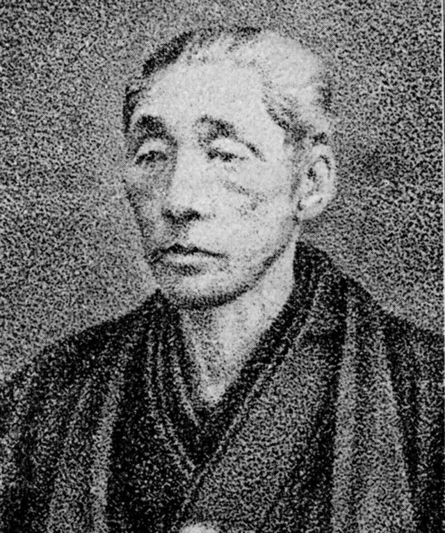 着物のような和装を身にまとい、落ち着いた表情で正面を見据える、白髪混じりの短髪で年配の男性の姿を捉えた、ザラつきのあるモノクロの質感が歴史的な背景を感じさせる石黒直寛(貫二)の肖像写真