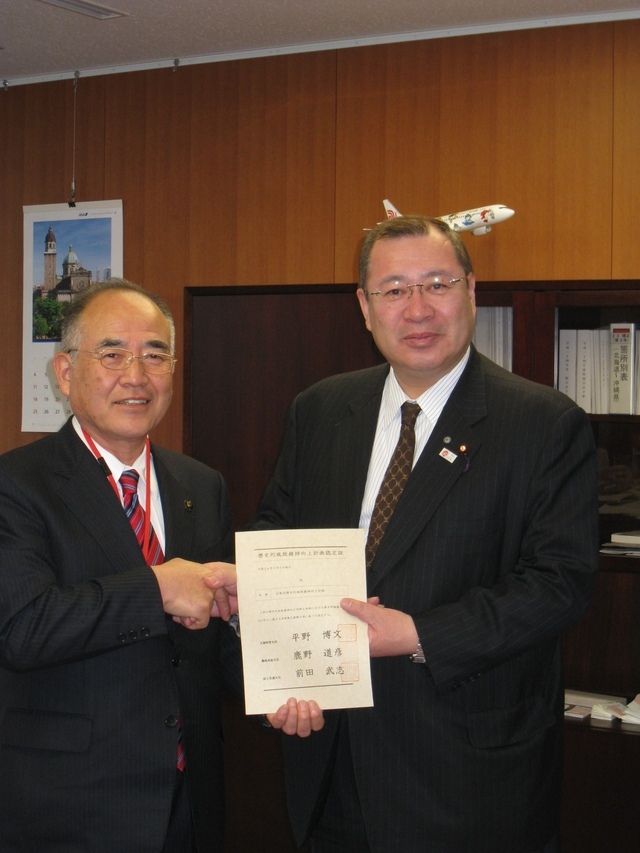 木目調の壁を背景に、スーツ姿の津島国土交通大臣政務官と秀島佐賀市長が「歴史的風致維持向上計画認定証」と記された書面を手に持ちながら、晴れやかな表情で握手を交わしている、認定式の厳かな一場面を捉えた写真