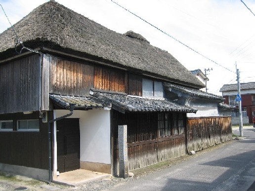 青空の下に佇む重厚な茅葺き屋根が特徴的な日本の伝統的な古民家で、経年変化した味わいのある木材の壁や瓦葺きの庇、白い漆喰壁が調和し、歴史の重みと当時の建築様式を今に伝える貴重な家屋の外観を捉えた写真