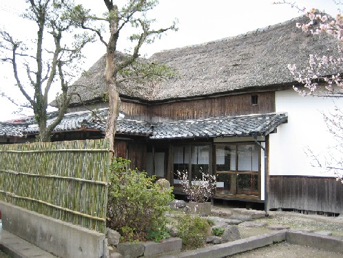 庭先に淡いピンク色の梅の花が咲き、竹垣に囲まれた趣のある日本庭園の奥に、重厚な茅葺き屋根と白い漆喰壁が美しい平屋建ての古民家が静かに佇んでいる写真