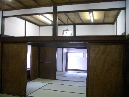 日本の伝統的な古民家の内部で、濃い茶色の太い梁や柱が白い漆喰壁と美しいコントラストを成し、開放された木製の引き戸の先には畳が敷き詰められた広々とした和室が奥まで続く、歴史を感じさせる静謐な室内風景の写真