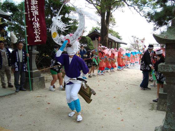 校庭のような開けた場所で、大きな角のような被り物をした踊り手が片足を高く上げ、その後ろでピンク色の衣装を纏い花笠を被った人々が列をなして伝統的な舞を披露している小松の浮立の写真