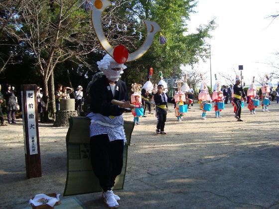 境内で、大きな鍬形のような被り物を着けた黒い衣装の踊り手が先頭に立ち、その後ろに色鮮やかな花笠を被り太鼓を抱えた子供たちが一列に並んで伝統的な祭りの儀式を行っている太田の浮立の写真