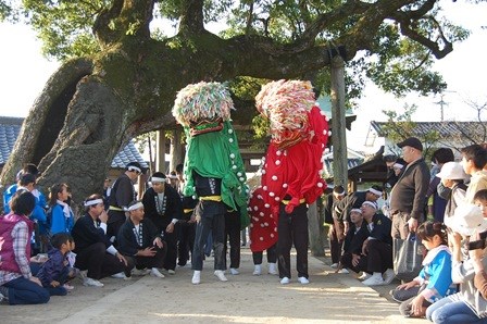 大きな幹に空洞がある立派な巨木の下で、緑と赤の鮮やかな獅子頭を被った演者が舞を披露し、法被姿の男性たちや近隣の住民たちがそれを取り囲んで見ている行事の風景写真