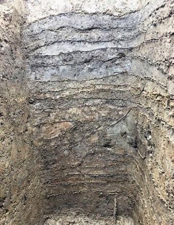 深く掘り下げられた地面の断面に、砂や粘土の層が幾重にも水平に積み重なり、強固に突き固められた街道の基礎構造が露出している様子を捉えた土層の写真。