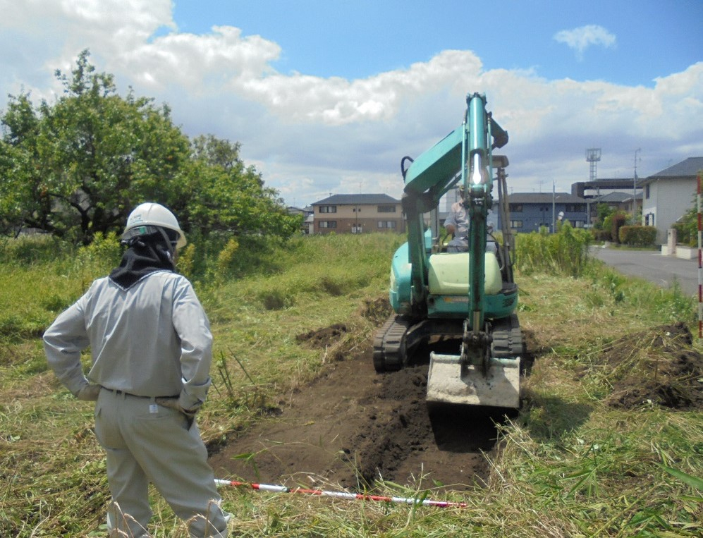 開けた草地にて、白いヘルメットと作業服を着用した人物が腰に手を当てて見守る中、エメラルドグリーンの小型ショベルカーが地面の土を掘り起こして整地作業を進めている発掘調査をしている様子の写真