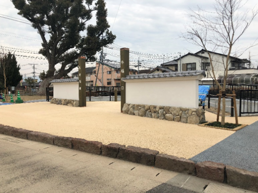明るいベージュ色の砂利が敷かれた敷地内に、石垣の土台と瓦屋根を備えた2枚の白い漆喰壁が並び、背後には大きな木や住宅街が広がる牛嶋口跡の入り口の写真