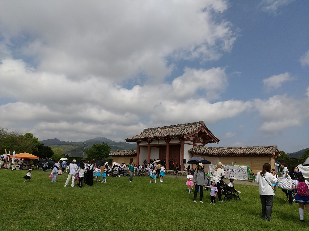 広々とした芝生広場に、復元された重厚な瓦屋根の古代門が佇み、その周辺でダンス衣装を纏った子供たちや家族連れが交流しているマルシェの様子の写真