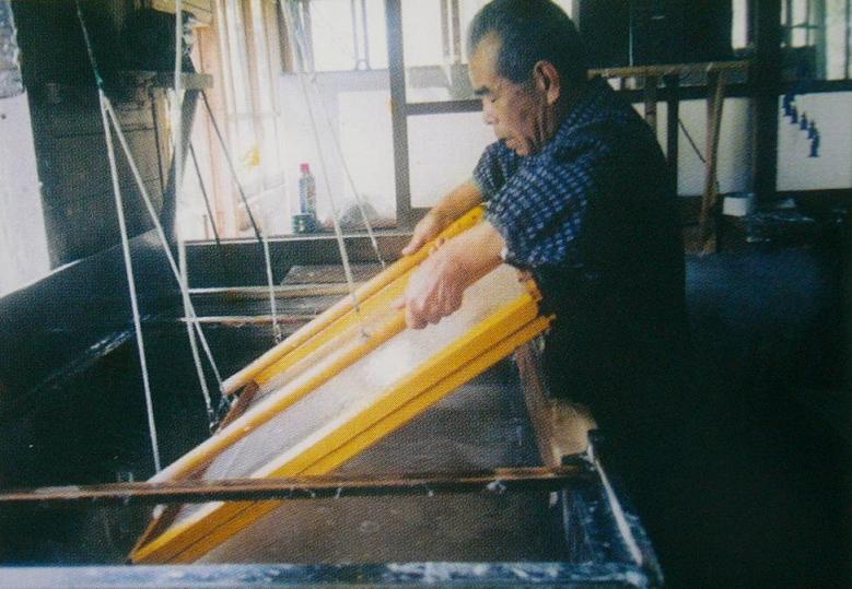 薄暗い工房の中で、男性が木製の大きな漉き枠を両手で持ち、水槽の中で原料を汲み上げながら一定のリズムで揺らして和紙を漉き上げている様子の写真