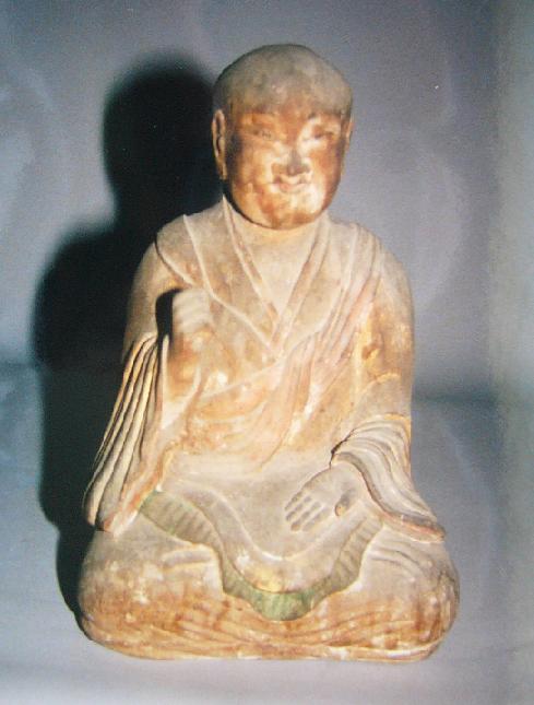 袈裟をまとった僧形の木彫像が両手を胸前と膝上に整えて静かに結跏趺坐し、柔らかな衣文の流れと落ち着いた佇まいが端正な造形美と重厚な存在感を示す写真