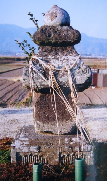 田園風景を背景に、注連縄が巻かれた古びた石造りの供養塔が静かに佇んでおり、頂部には宝珠が載り、台座の前には竹筒の献花台が置かれた石造宝篋印塔の写真