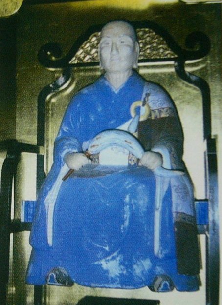 金色の背もたれを持つ椅子に青い衣をまとった坐像が巻物状の品を抱え静かに端正な姿勢で座す様子を収めた写真